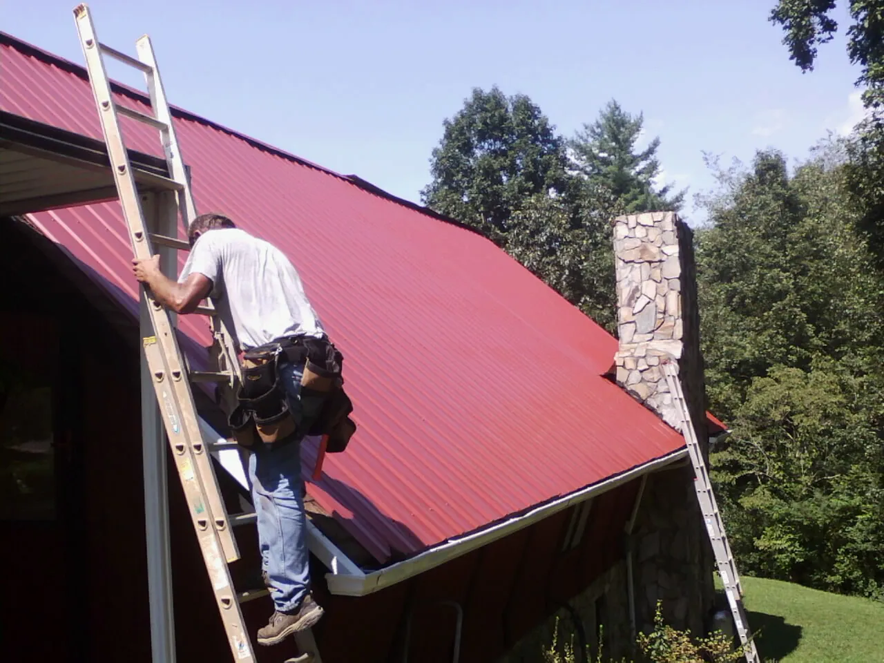 Roofer on a ladder beside a red metal roof and stone chimney — Soffit & Fascia Repair by Roofing Brothers in Bradenton Beach